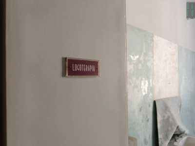 Proseguiamo il nostro giro. Sulle porte di alcune stanze si trova ancora la targhetta che ne indicava il reparto, come quella di colore rosso castagno che recita �logoterapia�